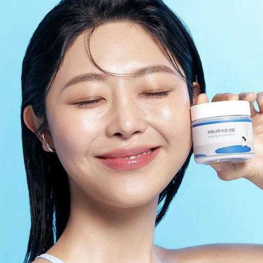 Femme souriant tenant sur sa joue le pot de crème ROUND LAB Birch Juice Moisturizing Cream sur fond bleu clair.