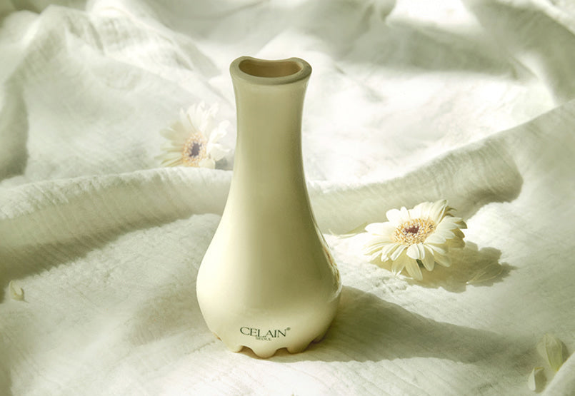 Gua Sha en forme de pot de fleur en céramique  avec base à picots texturés pour le massage stimulant et le drainage lymphatique. Sur un linge clair entouré de fleurs.