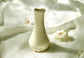 Gua Sha en forme de pot de fleur en céramique  avec base à picots texturés pour le massage stimulant et le drainage lymphatique. Sur un linge clair entouré de fleurs.