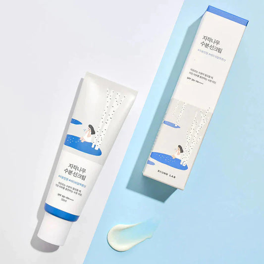 Tube de crème solaire Birch Juice Mosturizing Sunscreen de chez Round Lab Tube en plastique blanc, sur fond blanc. À sa droite la boîte du tube de crème solaire sur fond bleu clair. En bas de l’image une goutte de crème solaire.