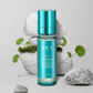 Brume Kira C-Tide Multi Mist de ZISU’C. Flacon transparent avec une étiquette bleu et capuchon bleu sur fond gris avec des galets et des plantes de cica.
